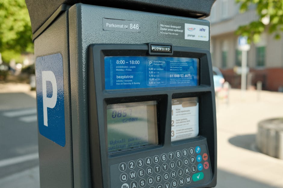 Close-up image of a modern parking meter situated outdoors on a paved street, with a blue parking sign displaying a white 'P' on the left side of the meter. The parking meter has a digital screen show